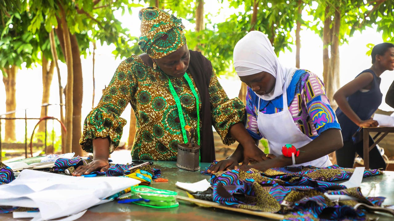 Zakaria Adama Lacera stands next to a young woman, a student, who is adjusting a piece of brightly coloured cloth that she has cut out.  A tape measure hangs around Zakaria’s neck. 