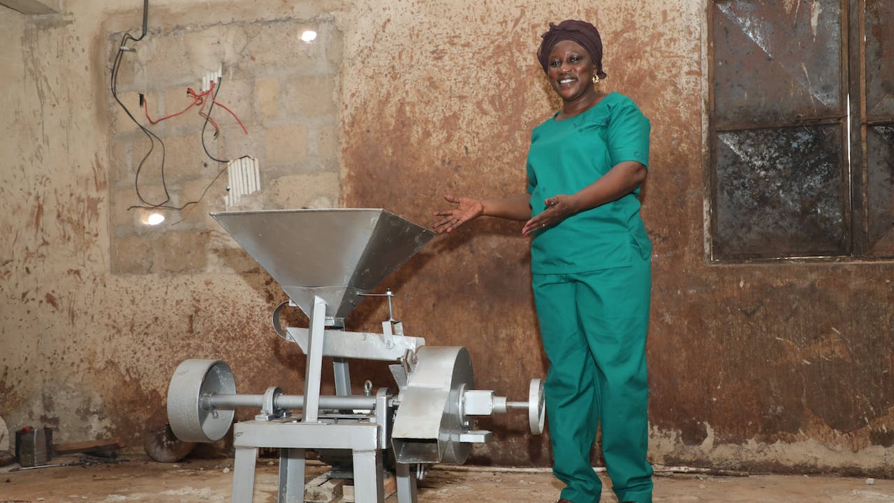 Zakaria Adama Lacera stands next to a new stainless steel machine and points towards it while smiling.